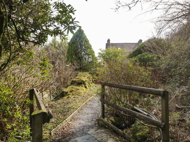 A gravel garden path bordered by mossy wooden railings and dense bushes with a stone building in the background at Duddon Cottage in Broughton-in-Furness