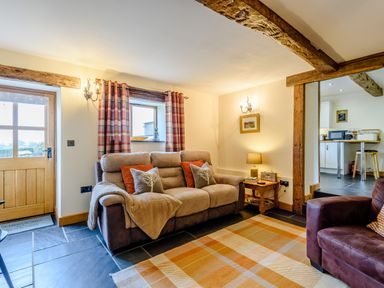 A living room with a sofa and armchair near a wooden door and window with plaid curtains at The Stables in Llanfair Caereinion near Welshpool
