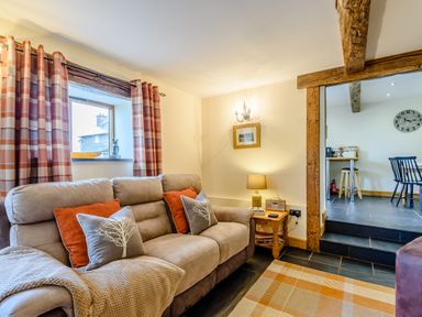 A living room with a beige sofa with cushions and a blanket next to a window with checkered curtains and an adjoining dining area at The Stables in Llanfair Caereinion near Welshpool
