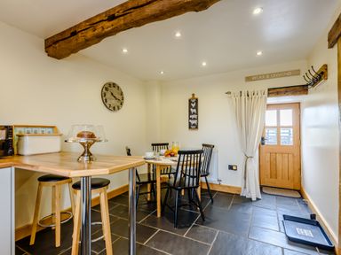 A kitchen dining area with a wooden breakfast bar and stools a small round dining table with four black chairs a wooden door with a curtain and a wall clock at The Stables in Llanfair Caereinion near Welshpool