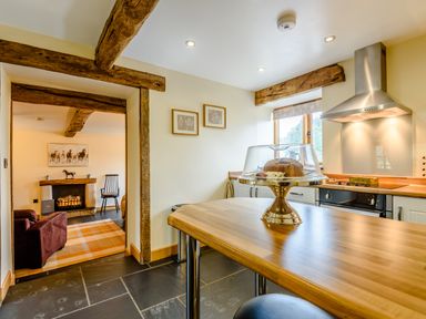 A kitchen with wooden beams and a wooden countertop with a cake stand overlooking a living room with a fireplace at The Stables in Llanfair Caereinion near Welshpool