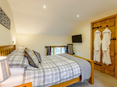 A bedroom with a wooden bed with plaid bedding two robes on a wooden door a wall-mounted television and a small window at The Stables Llanfair Caereinion near Welshpool