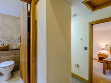 A hallway with a wooden door leading to a bathroom with a toilet and a bedroom with a bed and nightstand at The Stables in Llanfair Caereinion near Welshpool