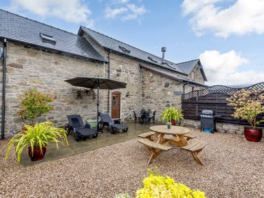 An outdoor patio with lounge chairs umbrella wooden picnic table plants and a barbecue at The Stables in Llanfair Caereinion near Welshpool