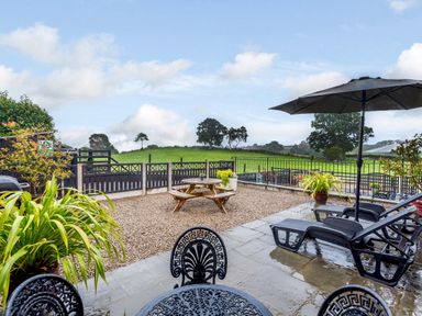 An outdoor patio with black chairs and tables and sun loungers with an umbrella overlooking a fenced garden and green fields at The Stables in Llanfair Caereinion near Welshpool
