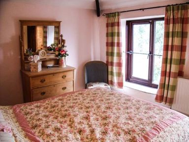 A bedroom with a wooden dresser with mirror flowers on top a chair by a window with checked curtains at Groom Cottage in Poughill near Bude