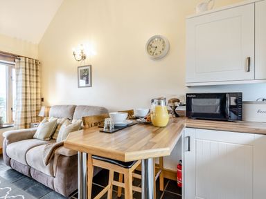 A kitchen area with a wooden countertop bar with stools bowls and juice next to a sofa with cushions and a window with curtains at The Granary in Llanfair Caereinion near Welshpool