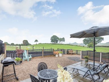 An outdoor patio with metal table chairs and umbrella and a fenced grassy field with trees at The Granary in Llanfair Caereinion near Welshpool
