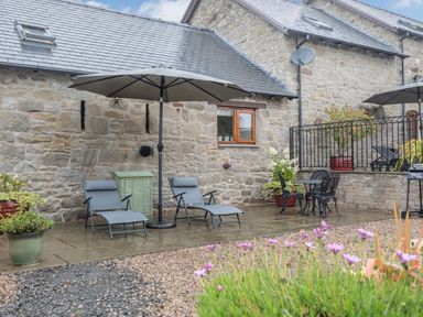 An outdoor patio area with two lounge chairs under an umbrella a small table with two chairs potted plants and a stone building at The Granary in Llanfair Caereinion near Welshpool