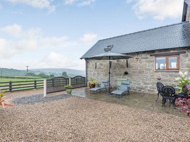 An outdoor patio area with two lounge chairs under an umbrella a stone wall a metal table and chairs and gravel ground at The Granary in Llanfair Caereinion near Welshpool