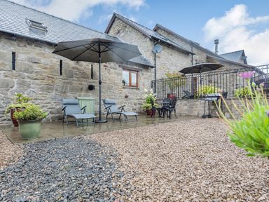 An outdoor patio area with two lounge chairs under an umbrella a small table with two chairs a barbecue grill and potted plants at The Granary in Llanfair Caereinion near Welshpool
