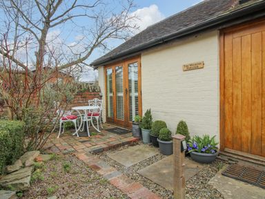An outdoor patio area with a small white metal table and two chairs with red cushions outside a building with wooden doors and potted plants at The Milking Parlour in Yarpole near Leominster