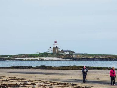 A sandy beach with two people walking near the shore and a lighthouse on a grassy island in the background at Sandy Knowes Low Hauxley near Amble