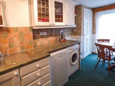 A kitchen with white cabinets dishwasher washing machine round wooden dining table and chairs at Sandy Knowes in Low Hauxley near Amble
