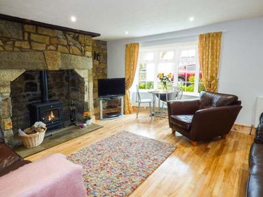 A living room with a stone fireplace wood burner a television leather armchairs a dining table with flowers and yellow curtains at Sandy Knowes in Low Hauxley near Amble