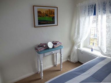 A bedroom corner with a small table holding folded striped towels and a plate near a window with lace curtains at Sandy Knowes in Low Hauxley near Amble
