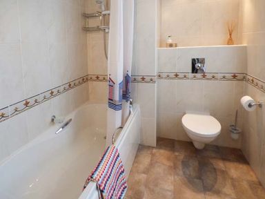 A bathroom with a bathtub a shower curtain a wall-mounted toilet and tiled walls at Sandy Knowes in Low Hauxley near Amble