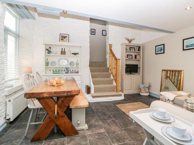 A dining and living area with a wooden table, chairs, a bookshelf, stairs, and a small TV at The Old Dairy in Maker near Millbrook