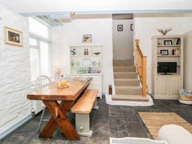 A dining area with a wooden table and bench next to stairs and a cabinet with a television at The Old Dairy in Maker near Millbrook