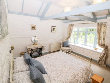 A bedroom with a bed, a chair by the window, a dressing table with a mirror, and light blue ceiling beams at The Old Dairy in Maker near Millbrook