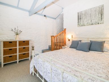 A bedroom with a bed with three pillows a basket chest and stairs against a white stone wall at The Old Dairy in Maker near Millbrook
