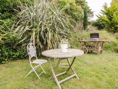 An outdoor garden area with a round wooden table and two metal chairs with a white potted plant on the table near tall plants and a wooden wheelbarrow with a barbecue grill on it at The Old Dairy Maker near Millbrook