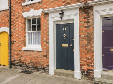 A brick building facade with a black door numbered 24 a window with white curtains and a yellow door at 24 College Lane Stratford-Upon-Avon