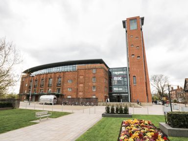 A large brick building with a tall tower and glass entrance with a welcome sign in a paved and grassy area with flowerbeds at 24 College Lane in Stratford-Upon-Avon