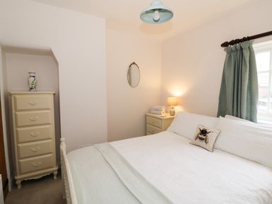 A bedroom with a bed with white bedding a nightstand with a lamp and towels a chest of drawers with a vase and a window with green curtains at 24 College Lane in Stratford-Upon-Avon