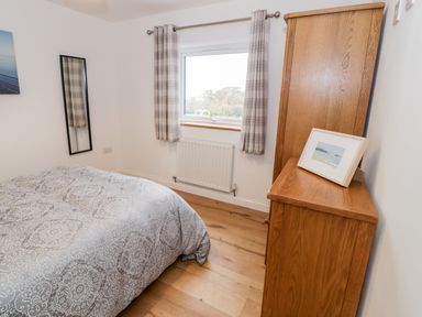 A bedroom with a bed covered by a patterned duvet a window with checkered curtains a wooden wardrobe and a framed picture on top at Ty'n Lon Bach in Llandwrog