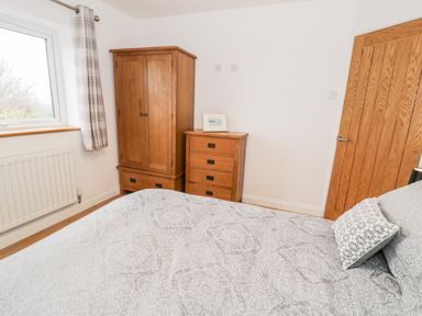 A bedroom with a wooden wardrobe, a chest of drawers, a bed with patterned bedding, a window with plaid curtains, and a wooden door at Ty'n Lon Bach in Llandwrog