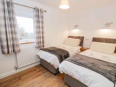 A bedroom with two single beds with white bedding and gray blankets two wall lamps and a window with plaid curtains at Ty'n Lon Bach in Llandwrog