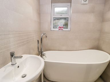 A bathroom with a freestanding white bathtub a white sink with a faucet and a window above the bathtub at Ty'n Lon Bach in Llandwrog