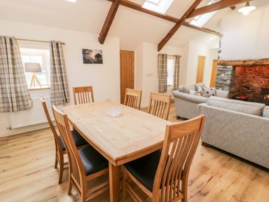A dining area with wooden table and six chairs beside a living room with grey sofas and a stone fireplace at Ty'n Lon Bach in Llandwrog