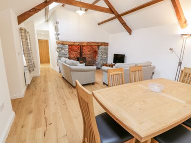 A living room with a wooden dining table and chairs two gray sofas a stone fireplace with a wood stove and exposed wooden ceiling beams at Ty'n Lon Bach in Llandwrog