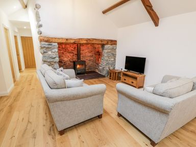 A living room with two grey sofas a wood-burning stove a TV on a wooden stand and wooden flooring at Ty'n Lon Bach in Llandwrog