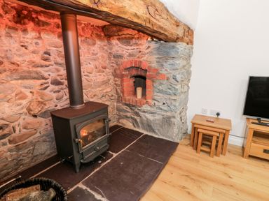 A stone wall with a wood-burning stove and candle in a niche along with nesting wooden tables and a television on a stand at Ty'n Lon Bach in Llandwrog