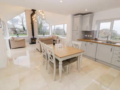A kitchen and dining area with a wooden table and white chairs next to a living room with beige sofas and a wood-burning stove at Ty'n Lon Bach in Llandwrog