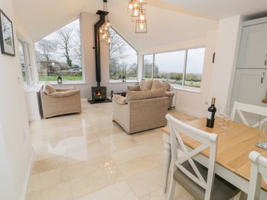 A sitting area with beige sofas and a wood stove surrounded by large windows next to a dining table with chairs and a bottle of wine at Ty'n Lon Bach in Llandwrog