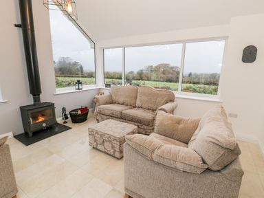 A living room with two sofas a wood stove a basket of firewood and large windows overlooking a field at Ty'n Lon Bach in Llandwrog