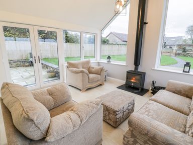 A sitting room with beige armchairs a floral patterned sofa an ottoman a wood burning stove and large windows at Ty'n Lon Bach in Llandwrog
