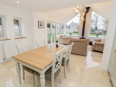 A dining area with a wooden table and four chairs next to a living room with two sofas and a wood stove at Ty'n Lon Bach in Llandwrog