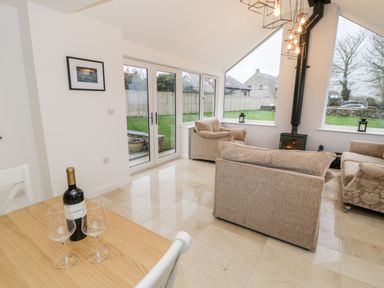 A living room with beige sofas a wood stove and large windows showing a garden at Ty'n Lon Bach in Llandwrog