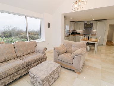 A living room with a sofa armchair and ottoman next to a window and a kitchen with dining table at Ty'n Lon Bach in Llandwrog