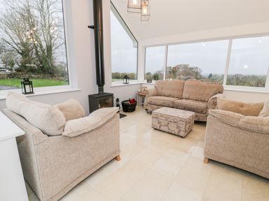 A living room with beige sofas a wood stove and large windows showing a garden and trees at Ty'n Lon Bach in Llandwrog