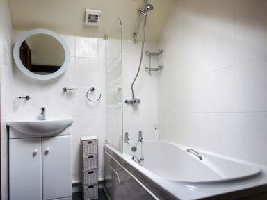 A bathroom with a bathtub shower combination a sink with cabinet a round mirror and a wicker storage unit at Kingfisher Barn in Staveley near Windermere