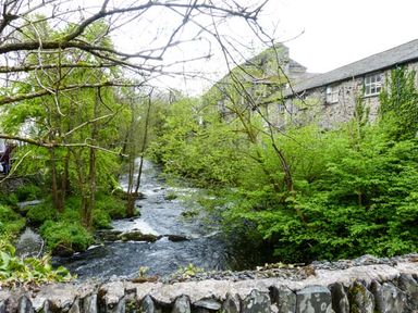 A flowing river surrounded by trees and a stone building behind a stone wall at Kingfisher Barn in Staveley near Windermere