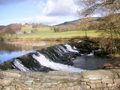 A stone wall in front of a river with a weir and grassy hills with trees in the background at Kingfisher Barn in Staveley near Windermere