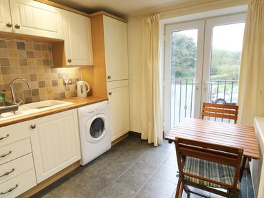 A kitchen with white cabinets washing machine wooden table two chairs and glass patio doors at Kingfisher Barn in Staveley near Windermere