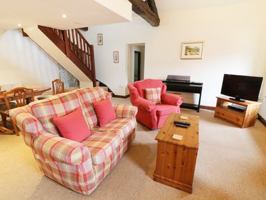 A living room with a plaid sofa and red armchair a wooden coffee table a TV on a wooden stand a piano and dining table near stairs at Kingfisher Barn in Staveley near Windermere